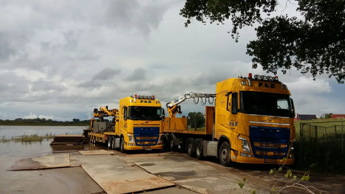 Twee P&K vrachtwagens lossen rijplaten bij een waterkant onder een bewolkte hemel.