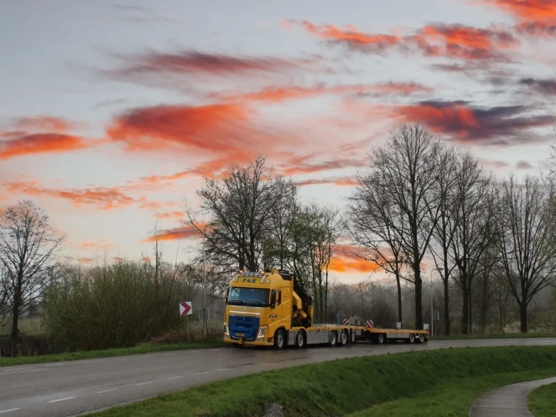 Sfeerfoto van een Autolaadkraan van P&K die op een weg rijdt met mooie oranje wolken op de achtergrond